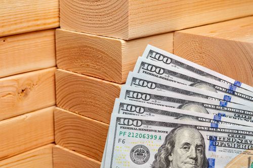 Why Do Lumber Prices Fluctuate? Stacks of 2x4s behind six $100 bills in United States currency.